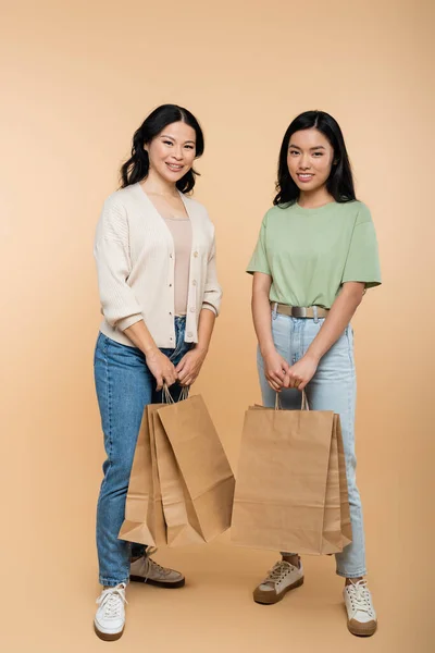 Volle Länge der glücklichen asiatischen Mutter und Tochter mit Papiertüten auf beige — Stockfoto