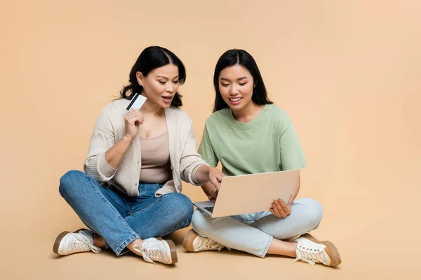 Asiatische Mutter und erwachsene Tochter mit Kreditkarte sitzen mit Laptop beim Online-Shopping auf beige — Stockfoto