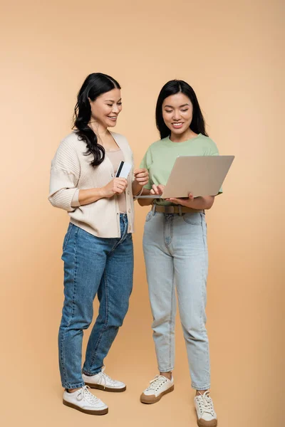 Happy asian mother and adult daughter with credit card looking at laptop during online shopping on beige — Stock Photo