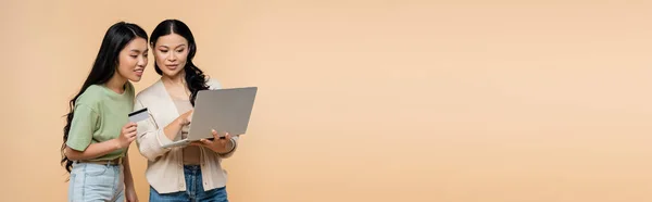 Asian mother and adult daughter with credit card looking at laptop during online shopping isolated on beige, banner — Stock Photo