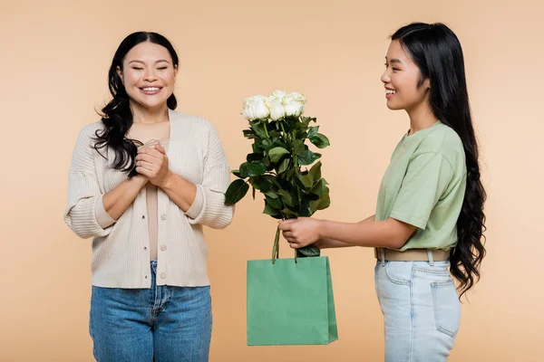 Happy asian daughter giving paper bag and flowers to smiling mother isolated on beige — Stock Photo
