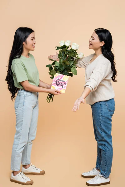 Happy asian daughter congratulating mother with greeting card and flowers on beige — Stock Photo