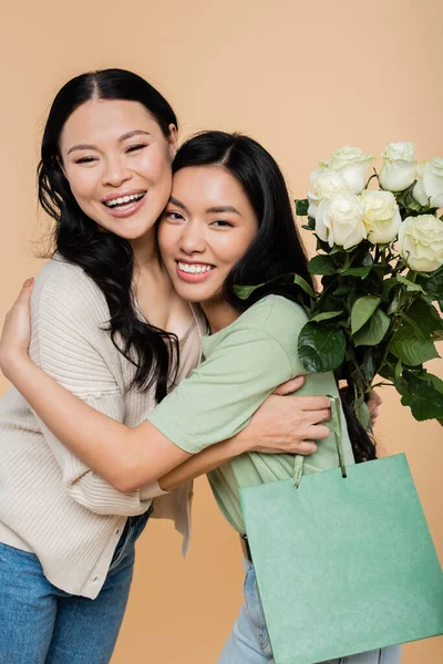 Happy asian mother holding flowers and hugging adult daughter isolated on beige — Stock Photo