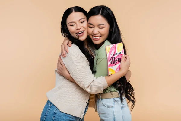 Asian mother holding greeting card and hugging happy adult daughter isolated on beige — Stock Photo