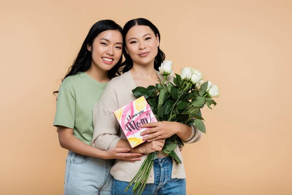 Glückliche asiatische Frau umarmt Mutter mit Blumen und Grußkarte isoliert auf beige — Stockfoto