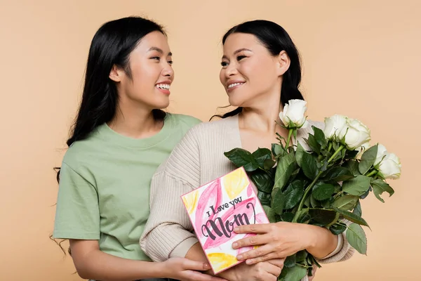 Cheerful asian woman hugging mother with roses and greeting card isolated on beige — Stock Photo
