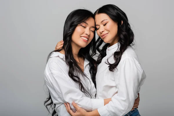 Joyful asian mother and young daughter in white shirts embracing isolated on grey — Stock Photo