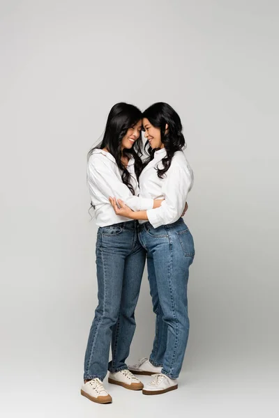 Longitud completa de alegre asiático madre y joven adulto hija en denim jeans y blanco camisas abrazando en gris - foto de stock