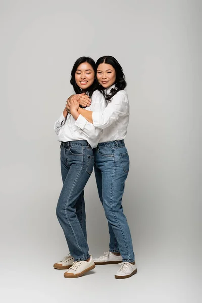 Full length of cheerful asian mother and daughter in denim jeans hugging on grey — Stock Photo