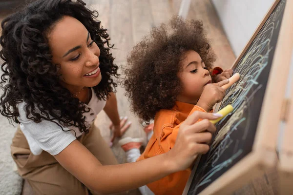 Vista ad alto angolo di donna afro-americana sorridente disegno sulla lavagna vicino figlia — Foto stock