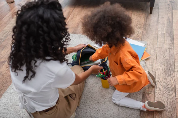 Vista ad alto angolo della madre afro-americana che tiene lo zaino vicino alla figlia con notebook a casa — Foto stock