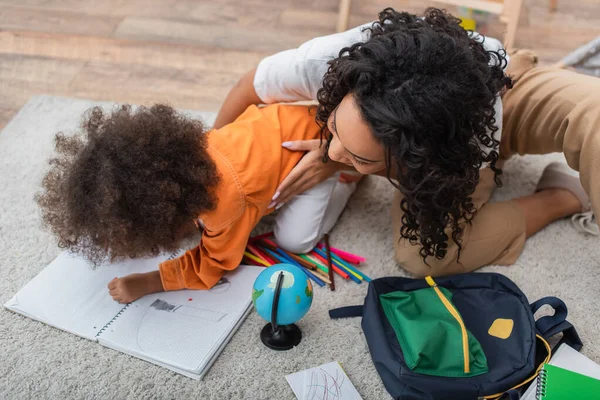 Madre afroamericana abrazando al niño dibujando en un cuaderno cerca del globo y la mochila - foto de stock