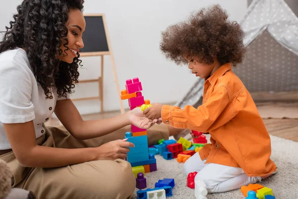 Vista lateral de la sonriente mujer afroamericana jugando bloques de construcción cerca de su hija en casa - foto de stock