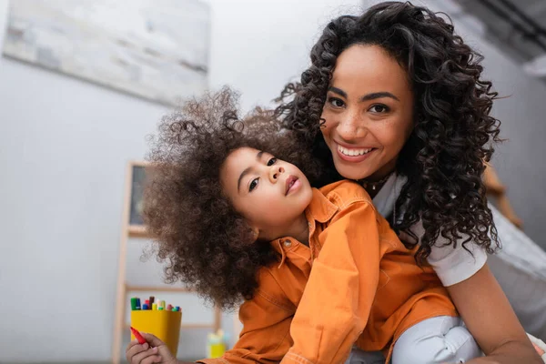 Mãe afro-americana positiva olhando para a câmera perto da criança e lápis de cor em casa — Fotografia de Stock