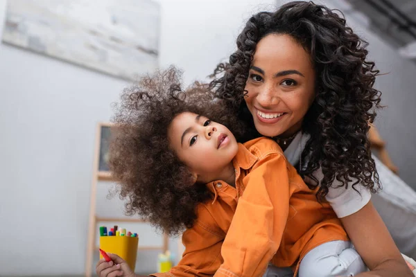 Positiva madre afroamericana mirando a la cámara cerca de niños y lápices de color en casa - foto de stock