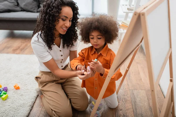 African american mother holding chalks near daughter, chalkboard and building blocks — Stock Photo