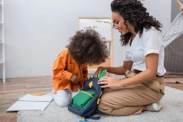 Feliz afroamericana mujer poniendo portátil en la mochila cerca de niño en casa - foto de stock