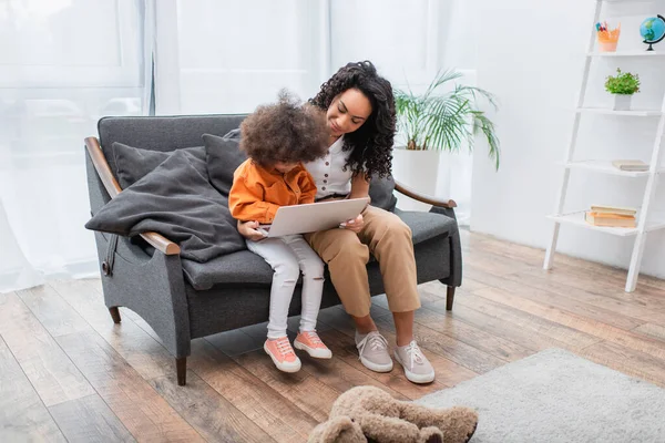 Sonriente mujer afroamericana sosteniendo portátil cerca de niño en el sofá - foto de stock