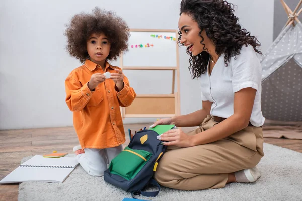 Glückliche afrikanisch-amerikanische Mutter betrachtet Tochter in der Nähe von Notizbüchern und Rucksack im Wohnzimmer — Stockfoto