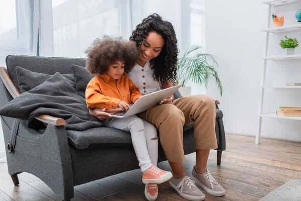 Mère afro-américaine utilisant un ordinateur portable près de l'enfant sur le canapé dans le salon — Photo de stock