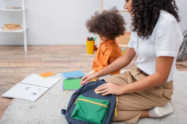 Niño afroamericano sosteniendo lápices de color cerca de cuadernos y madre con mochila en casa - foto de stock