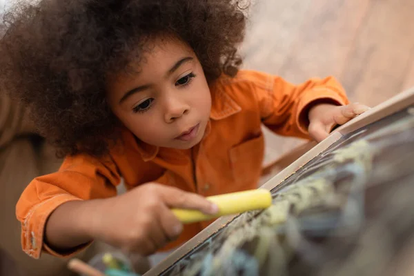 Draufsicht eines afrikanisch-amerikanischen Kindes, das zu Hause auf unscharfer Kreidetafel zeichnet — Stockfoto