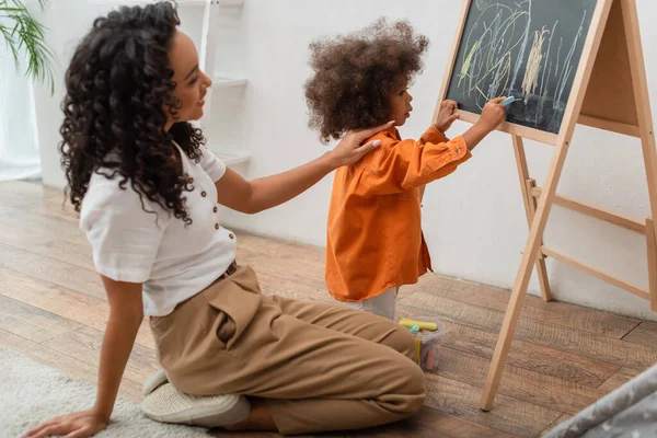 Bambino bambino disegno sulla lavagna vicino sorridente madre afro-americana a casa — Foto stock