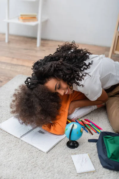 Heureux parent afro-américain étreignant enfant en bas âge près du globe, cahiers et crayons de couleur à la maison — Photo de stock