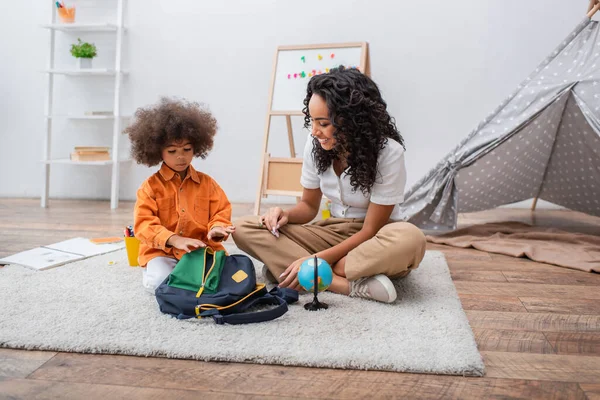 Enfant tout-petit assis près du globe, cahiers et maman afro-américaine dans le salon — Photo de stock