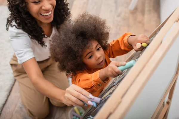 Draufsicht der lächelnden afrikanisch-amerikanischen Mutter, die auf Kreidetafel neben Kleinkind zu Hause zeichnet — Stockfoto