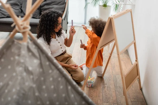 Toddler african american kid pointing at chalk near mom and tent at home — Stock Photo