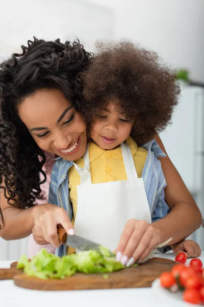 Lächelnde afrikanisch-amerikanische Mutter schneidet Salat neben Tochter und Kirschtomaten in Küche — Stockfoto