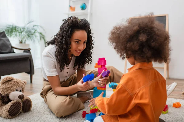 Joven afroamericana mujer sosteniendo bloques de construcción cerca de hija y osito de peluche en casa - foto de stock