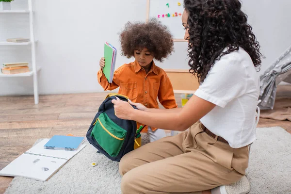 Criança afro-americana segurando notebook perto da mãe com mochila na sala de estar — Fotografia de Stock