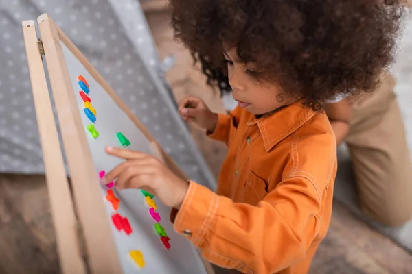 Vista de ángulo alto del niño afroamericano jugando con carteles en el tablero magnético cerca de mamá en casa - foto de stock