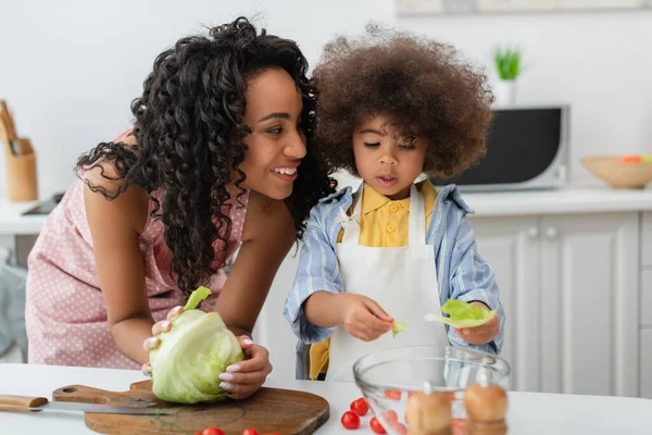Улыбающаяся африканская американка держит капусту рядом с дочерью в фартуке на кухне — стоковое фото