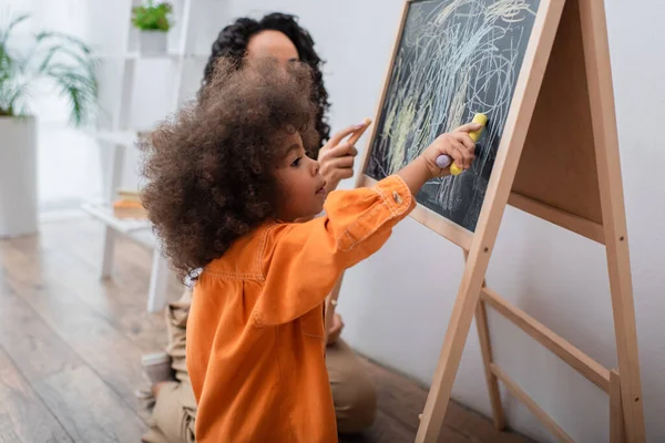 Kleinkind afrikanisch-amerikanische Mädchen zeichnen auf Kreidetafel in der Nähe von Mama zu Hause — Stockfoto