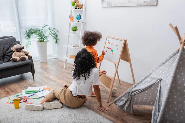 Africaine américaine fille debout près du tableau magnétique, mère et tipi à la maison — Photo de stock