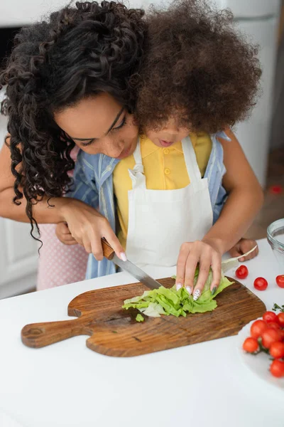 Donna afroamericana che taglia lattuga vicino al bambino in grembiule in cucina — Foto stock