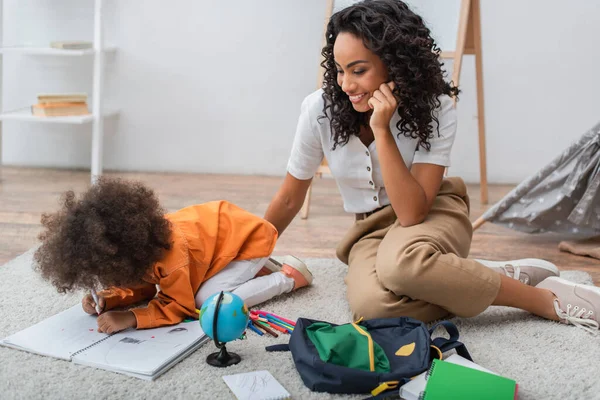 Sorridente donna africana americana seduta vicino alla figlia del bambino che disegna sul taccuino a casa — Foto stock