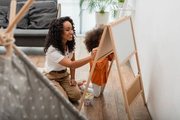 African american parent and daughter drawing on chalkboard near tent at home — Stock Photo