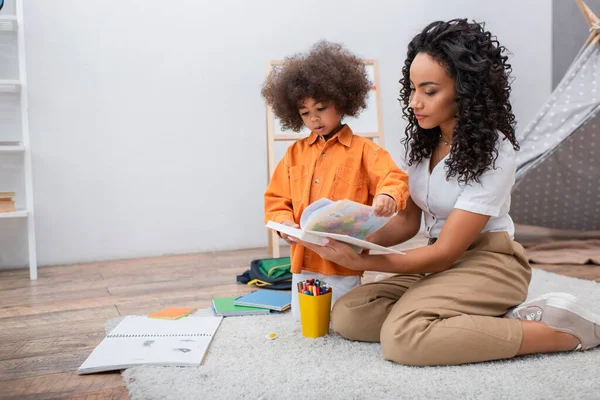 African american mother and daughter holding book near color pencils and notebooks at home — Stock Photo