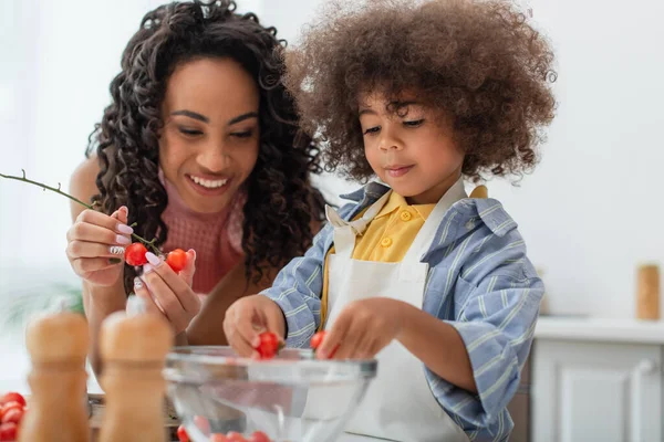 Enfant afro-américain tout-petit tenant des tomates cerises près de maman souriante dans la cuisine — Photo de stock