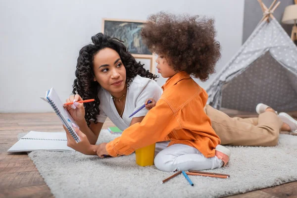 Mujer afroamericana sosteniendo cuaderno cerca de la hija y lápices de color en la alfombra en casa - foto de stock
