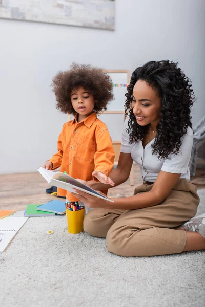 Femme afro-américaine positive tenant un livre près de sa fille et des crayons de couleur à la maison — Photo de stock