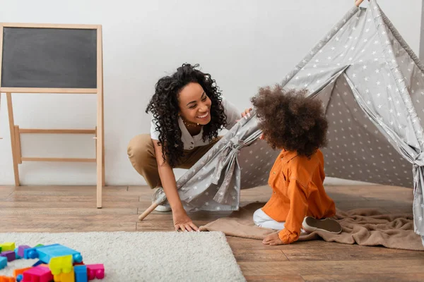 Sonriente madre afroamericana mirando al niño en tipi en casa - foto de stock