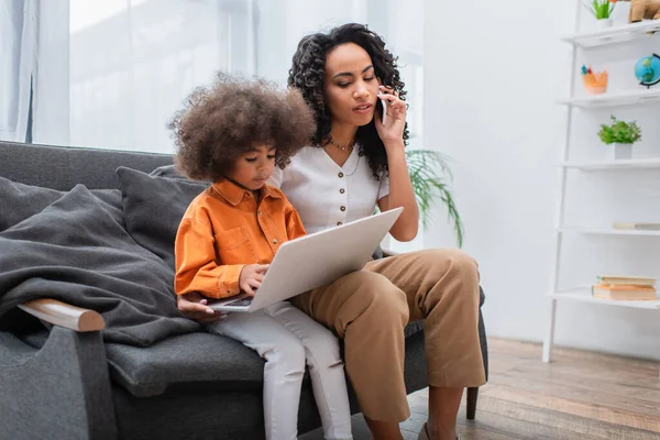 Mujer afroamericana hablando en un teléfono inteligente cerca de su hija utilizando el ordenador portátil en el sofá - foto de stock