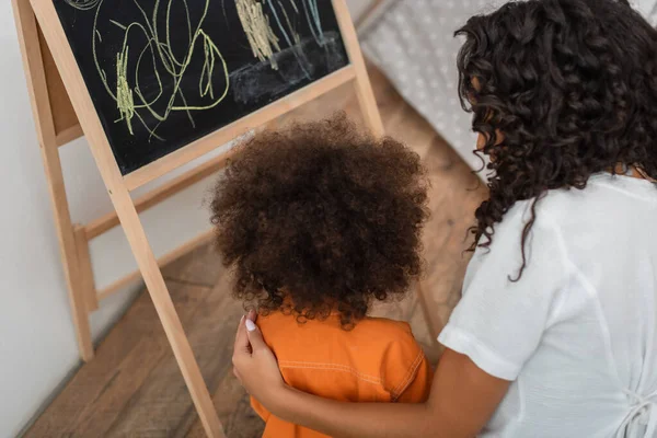 Vista aérea de una mujer afroamericana abrazando a un niño cerca de la pizarra en casa - foto de stock
