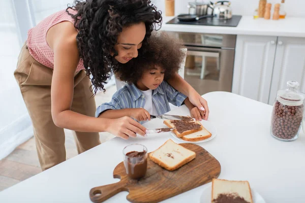 Sonriente africano americano mamá difusión chocolate pasta en pan cerca hija en cocina - foto de stock