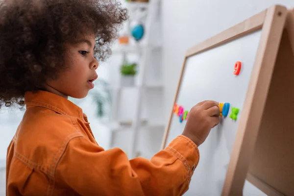 Vue latérale de l'enfant afro-américain appliquant des signes sur le tableau magnétique à la maison — Photo de stock